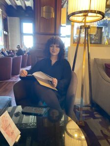 Photo of Alex M, sitting in a chair holding a book. There is a lamp to the right and a table in the foreground.