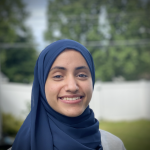 Headshot of Rogayah in a blue hijab, standing in front of trees