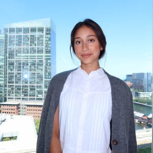 Photo of Sophie in front of the Atlantic Wharf in Boston. She is wearing a white top and gray sweater.
